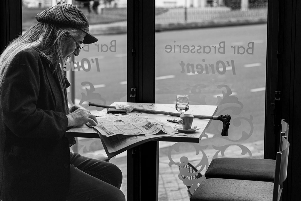 Un homme âgé aux cheveux longs avec une casquette dans un café est en train de lire un journal